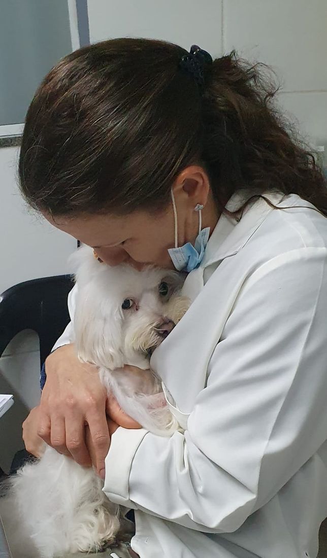 Dra. Maria Angélica, de jaleco, acolhendo com a testa um cãozinho branco em seu consultório
